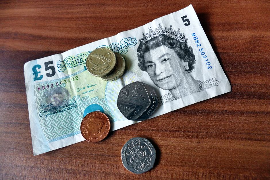 A crumpled five-pound banknote featuring a portrait of Queen Elizabeth II is spread on a wooden surface, alongside various UK coins including pennies, two-pound, ten-pence, and one-pound pieces. The b