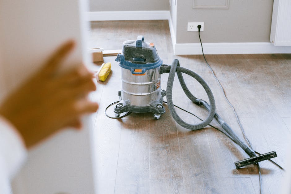 A person kneeling on a patterned carpeted floor, engaging with a yellow portable vacuum cleaner installed with a vacuum hose. The individual is wearing a beige jacket and blue jeans, with their head a
