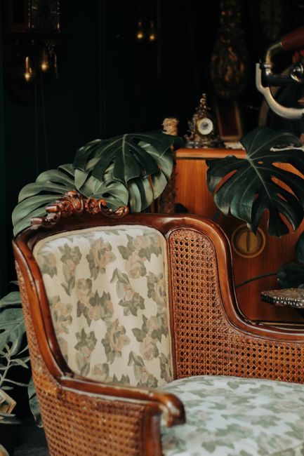 A close-up view of an antique armchair with a wooden frame and ornate gold detailing, upholstered in a cream-colored fabric with a subtle pattern. The chair is positioned in a room with wooden floorin